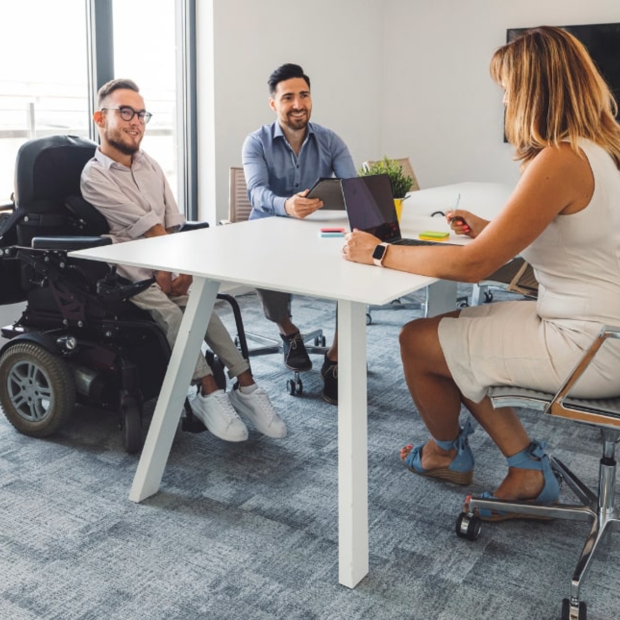 A woman with shoulder length blond hair wearing a white dress sits with her back to us at a desk in an office. Sat across the desk are two white men; one is a wheelchair user wearing a light shirt and glasses, the other is non-disabled with dark hair